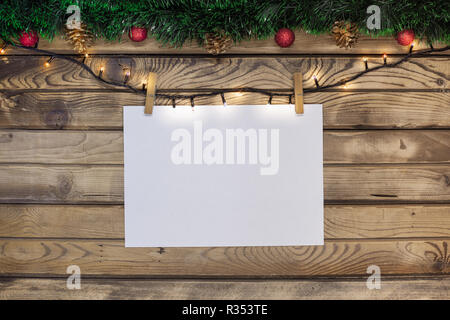 Gruß an Weihnachten auf einem weißen Blatt hing an einen Kranz mit Wäscheklammern auf einem hölzernen Hintergrund mit grünem Lametta und rot Spielzeug eingerichtet Stockfoto