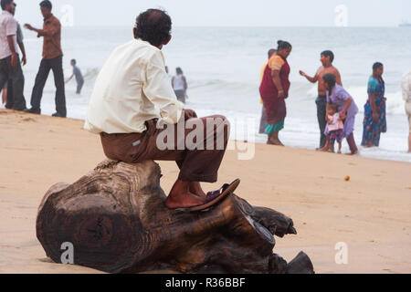 Chennai, Indien - 6. September 2007: ein Mann sitzt auf einem Baumstumpf Blicke in Richtung der rauen See, wie andere Leute amüsieren sich im Marina Beach in Chen Stockfoto