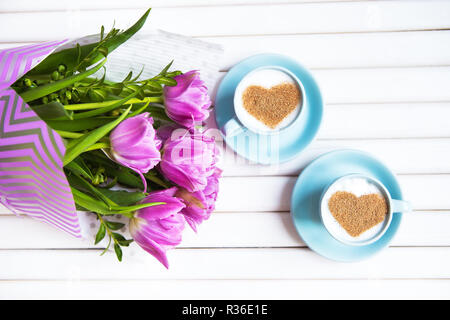 Zwei Tassen Cappuccino mit einem Herzförmigen Symbol und lila Tulpen auf einer hölzernen Hintergrund. Stockfoto