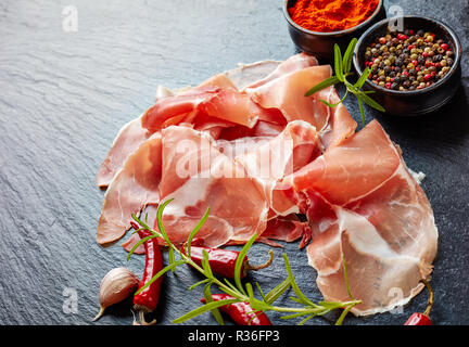 Spanisch trocken - Schinken, jamon Curado dünn geschnitten und serviert auf einem schwarzen Stein Platte mit Rosmarin und Arten, horizontale Ansicht von oben, close-up Stockfoto