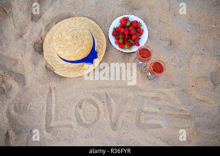 Strohhut, Wein und einen Teller mit Erdbeeren auf einem Sandstrand - aufschrift Liebe. Stockfoto