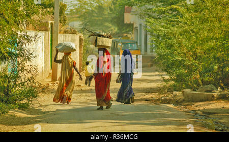 RAJASTHAN INDIEN staubige Straße und drei Frauen mit bunten Saris und SCHWEREN LASTEN AUF DEM KOPF Stockfoto