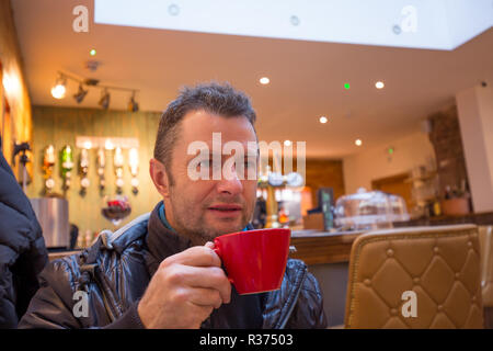 Nahaufnahme eines attraktiven kaukasischen Männchens, Mitte vierziger, in der trendigen Bar, bei einem Kaffee aus der roten Tasse, die er hält. Stockfoto