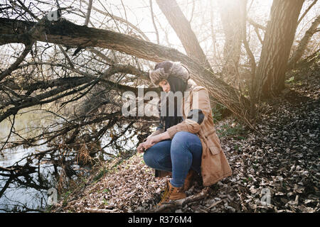 Junge brünette Frau kauerte am Ufer eines Flusses trägt eine Jacke und eine Russische Bomber leder hut mit dem Boden voller Blätter im Herbst Stockfoto