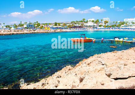 Bild von einem roten Schnellboot in der Bucht von vathia Gonia Beach in der Nähe von Agia Napa, Zypern. Felsige Küste und Meer mit türkisblauem Wasser in einer Bucht, Häuser auf Stockfoto