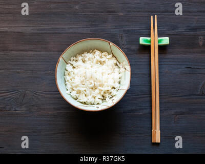 Blick von oben auf die Portion gekochtem Reis in der Schüssel und Essstäbchen auf Rest auf dunklem Holzschreibtisch Stockfoto