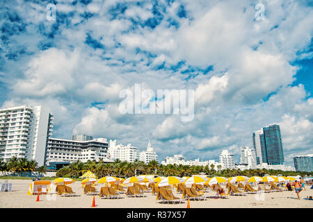 Miami, South Beach, der Strand mit Liegestühlen unter der gelben Sonnenschirmen, grüne Palmen und Mehrfamilienhäuser, Hotels oder sonnigen Tag. Die Skyline der Stadt mit weißen Wolken am blauen Himmel. Sommer Urlaub. Städtische Landschaft Stockfoto