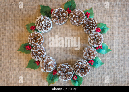 Weihnachten Kranz von Tannenzapfen, Holly Blättern und roten Beeren auf rustikalen Stoff Hintergrund Stockfoto