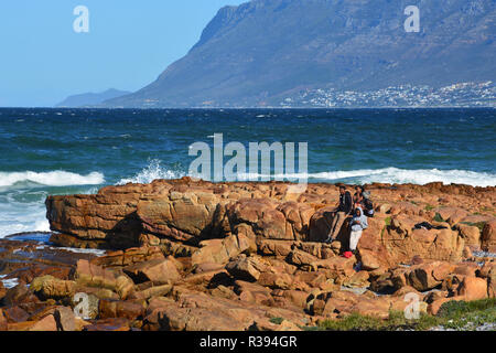 Eine Gruppe von Jugendlichen heraus hängen auf den Felsen an der Küste der False Bay zwischen Strand von Muizenberg und Fish Hoek außerhalb von Kapstadt, Südafrika Stockfoto