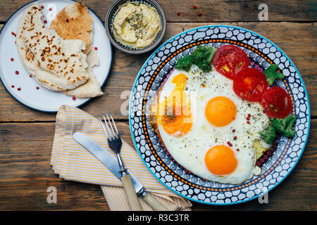 Frühstück Tabelle einstellen. Spiegeleier auf einem Teller mit Tomaten, Brot (Pita) und Hummus auf einem Holztisch. Ansicht von oben. Holz- Hintergrund. Stockfoto