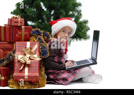 Kleines Mädchen in Santa Claus Cap mit einem Laptop und Weihnachten Geschenke auf weißem Hintergrund, isoliert Stockfoto