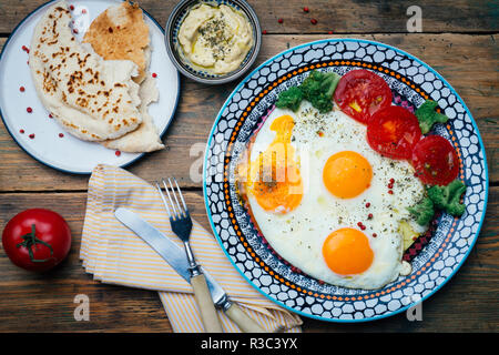 Frühstück Tabelle einstellen. Spiegeleier auf einem Teller mit Tomaten, Brot (Pita) und Hummus auf einem Holztisch. Ansicht von oben. Holz- Hintergrund. Stockfoto