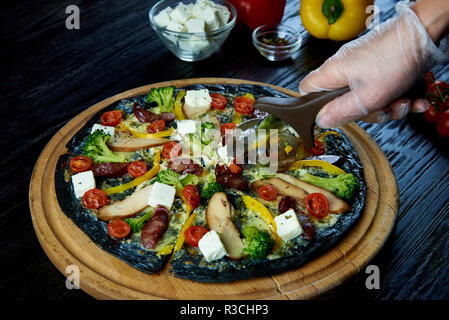 Pizza in Scheiben schneiden mit einem speziellen Messer. Frisches Gemüse, Käse und Gewürze im Hintergrund. Hand in Hand mit einem Messer. Stockfoto