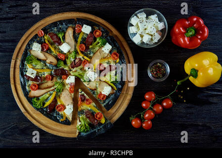 Hot aromatisiert köstliche italienische Pizza auf schwarz Teig mit Fleisch, Gemüse und Käse auf einem dunklen Holzplatte mit rohen, Paprika, Tomaten, Käse ein Stockfoto