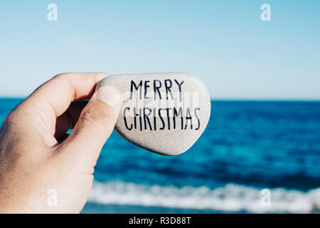 Nahaufnahme von einem jungen Mann mit einem Stein in der Hand, mit dem Text frohe Weihnachten handschriftlich in es, am Strand, mit dem Meer im Hintergrund Stockfoto