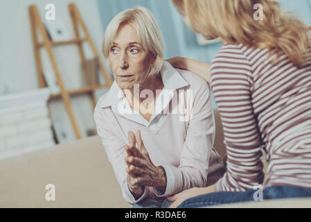 Melancholie. Portrait von traurige ältere Frau Palmen zusammen zu halten und nachdenklich auf der Suche auf der linken Seite der Kamera, während jungen Tochter saß in der Nähe und Stockfoto