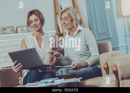 Angenehme Dialog. Liebenswerte ältere Dame mit Notizblock in die Hände am Laptop Bildschirm junger erfreut, Frau neben ihr sitzt, beim Besprechen Stockfoto