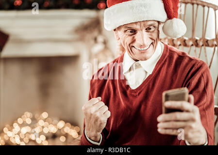 Ich habe es geschafft. Zufrieden überrascht alter Mann verbringt Zeit im hellen Zimmer Jubel und sein Handy halten. Stockfoto