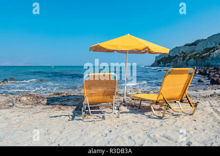 Zwei gelbe Stühle und ein gelbes Dach stehend vor dem azurblauen Mittelmeer an einem weißen Sandstrand in Griechenland. Stockfoto