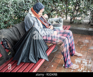 Barfuß Obdachloser sitzt auf der Bank im Regen mit Fuß in der Pfütze. Stockfoto