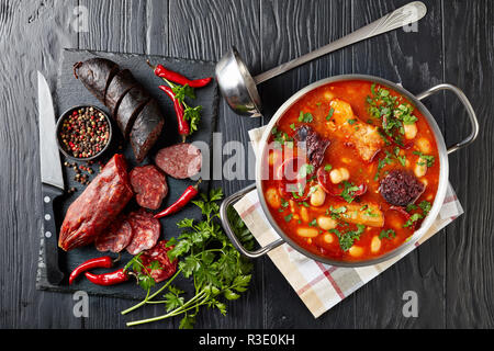 Herzhafte Fabada Asturiana Bohneneintopf mit Chorizo, Speck und Blut Wurst in eine Kasserolle aus Metall auf einem schwarzen Holztisch mit Zutaten auf einem Schneiden bo Stockfoto