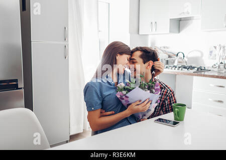 Junger Mann gifting Blumenstrauß zu seiner Freundin in der Küche. Glückliches Paar umarmte. Romantische Überraschung zu Hause Stockfoto
