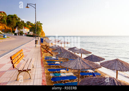 Holzbank, auf Fliesen- Gehweg hinter, aber über dem öffentlichen Strand mit strohgedeckten Sonnenschirmen, Sonnenschirmen und Liegen neben der Küste. Stockfoto