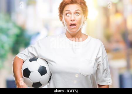 Attraktives senior kaukasischen rothaarige Frau mit Fußball über isolierte Hintergrund im Schlag mit einer Überraschung konfrontiert, ängstlich und aufgeregt mit f Angst Stockfoto