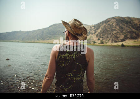 Ehrliches Foto eines Mannes mit Cowboyhut, der einen Blick auf den Fluss hat und die Aussicht genießt. Fläche in nicht sichtbar. Stockfoto