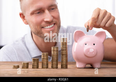 Glückliche junge Mann einfügen Münze in Sparschwein in der Nähe von steigenden Gewinn auf Schreibtisch Stockfoto