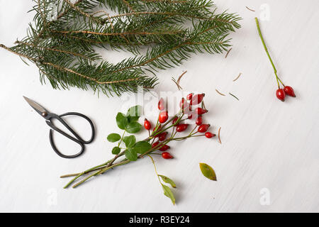 Vorbereitung natürlichen Weihnachtsdekoration mit Hagebutten, Tannenzweigen und Schere auf einem weißen Tisch mit Kopie Raum, hohe Engel Blick von oben, Stockfoto