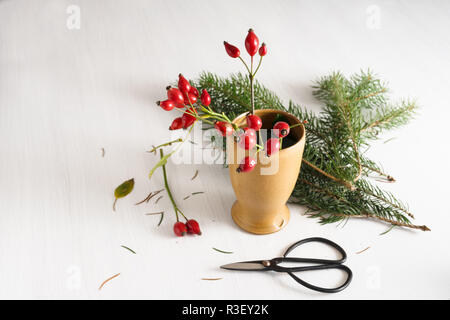 Vorbereitung eines natürlichen Weihnachtsdekoration in einem Steinzeug Vase mit Hagebutten, Tannenzweigen und Schere auf einem weißen Tisch mit Kopie Raum, Stockfoto