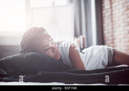 Eine schöne afrikanische Frau liegt und schläft im Bett, in einem weißen Schlafzimmer. Guten Morgen, Urlaub zu Hause. Das Mädchen trägt ein T-Shirt, Pigtails auf dem Kopf Stockfoto