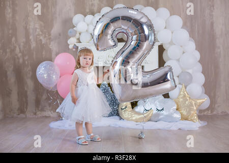 Zwei Jahr Baby Mädchen in weiß rosa Kleid in Studio shoot posiert mit großen riesigen Silber Ballon Nummer 2 und viel Weiß luftig baloons. Stockfoto