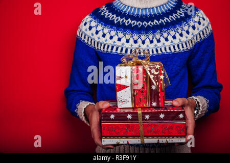 Studio geschossen von einem jungen Mann in Isländische pullover Holding ein Haufen Geschenkboxen auf rotem Hintergrund. Weihnachten oder Silvester feiern. Stockfoto