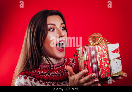 Studio geschossen von einer jungen Frau, die in der isländischen Pullover Holding eine Geschenkverpackung auf rotem Hintergrund. Weihnachten oder Silvester feiern. Stockfoto