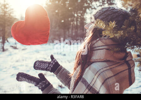 Junge Frau wirft einen roten Kissen Herz im Winter Wald bei Sonnenuntergang Stockfoto