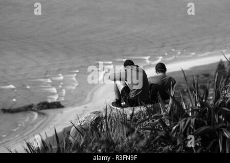 Zwei Freunde sitzen auf einem Berg mit Blick auf das Meer am Mount Maunganui, Neuseeland Stockfoto