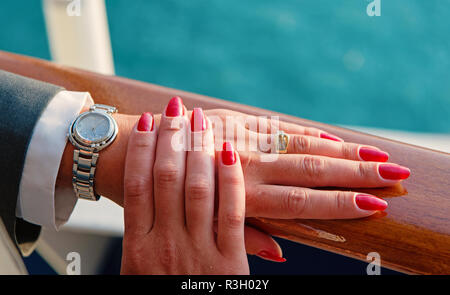 Weibliche Hände und Armbanduhr mit roten Maniküre und goldenen Ring am Finger mit Diamanten in der Nähe von Wasser, schöne Hände mit ansehen. Stockfoto