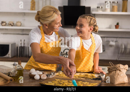 Gerne Großmutter und Enkelin in Schürzen lächelnd während sie cookies zusammen vorbereiten Stockfoto