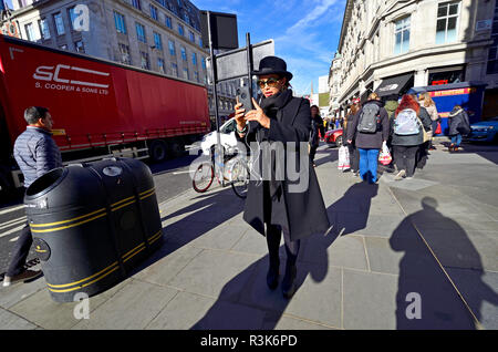 London, England, UK. Frau mit Hut, nehmen auf Ihr Handy beim Gehen Stockfoto