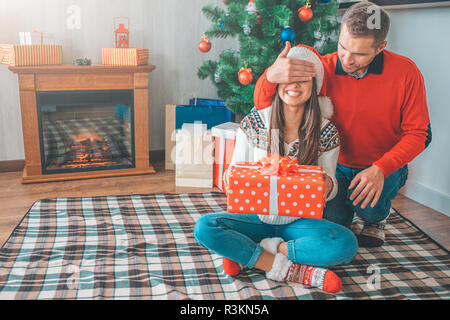 Schönes Bild des Menschen in der Gruppe Position hinter Frau sitzt. Er hält Ihre Augen mit der Hand und schaut sie geschlossen. Sie lächelt und hält Big Red Box mit Stockfoto