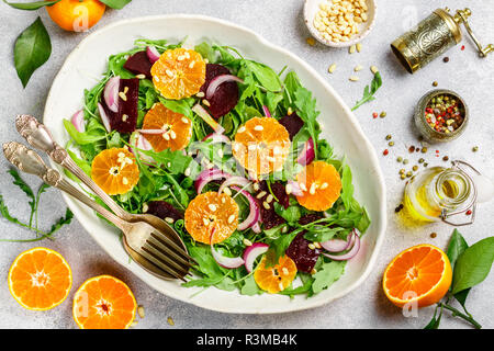 Feinkost bunter Salat mit gebackenen Rüben, Rucola, Mandarinen und roten Zwiebeln mit Gewürzen und Pinienkerne in einer weißen Schüssel. Olivenöl und Zutaten auf Stockfoto