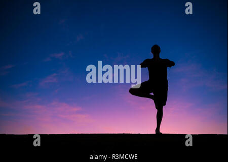 Silhouette des Menschen auf ein Bein in einem ständigen yoga Baum Posieren vor der farbenfrohen Sonnenaufgang Himmel Stockfoto