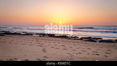 Fußspuren im Sand führen in Richtung Ozean während des Sonnenuntergangs. Clear Sky. Goldene Stunde. Kopieren Sie Platz. Stockfoto