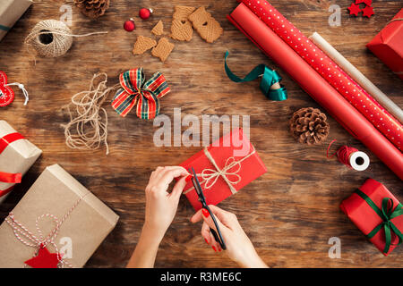 DIY Geschenkverpackung. Frau Verpackung schöne rote Weihnachtsgeschenke auf rustikalen Holztisch. Overhead Sicht von Weihnachten folierstation. Stockfoto