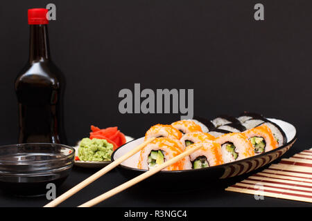 Uramaki Kalifornien. Sushi Rollen mit Nori, Reis, Avocado, Gurke, mit fliegenden Fischen roe auf Keramik Platte eingerichtet. Flasche und Schüssel mit so Stockfoto