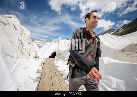 Glaciolog David Volken an der Mündung des Rhone-Gletschers. Riesige Laken mit Fleece-Decken, die einen Eistunnel an der Mündung des Rhonegletschers in der Schweiz bedecken. Nach einem Winter mit Rekordschneemengen war der größte Teil davon verschwunden, als dieser Imae am 14. Juli 2018 aufgenommen wurde, um das dunklere Eis freizulegen. Während Schnee ein brillanter Reflektor der Sonnenenergie ist, absorbiert das dunklere Eis stattdessen die Energie und beschleunigt so das Schmelzen des Gletschers. Die Farbe und Dunkelheit des Gletschereises variiert weltweit, je nach Verschmutzungsbildung, Alter des Eises, vom Eis aufgepflückten Partikeln und von Stockfoto