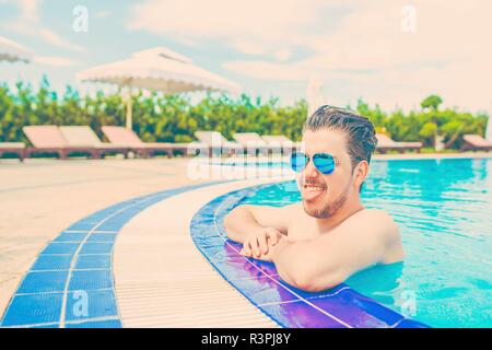 Man schaut aus dem Pool, hängen an der Reling. Junger Mann, eine gute Zeit im Pool und zeigt Zunge, quält. Blaugrün und orange Tönen. Stockfoto