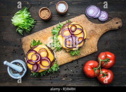 Zwei leckere saftige home Burger mit Rindfleisch, Salat, eingelegte Gurken, Käse, Zwiebeln, Tomaten, Brötchen Mit Sesam, auf einem vintage Schneidbrett, oben vi. Stockfoto